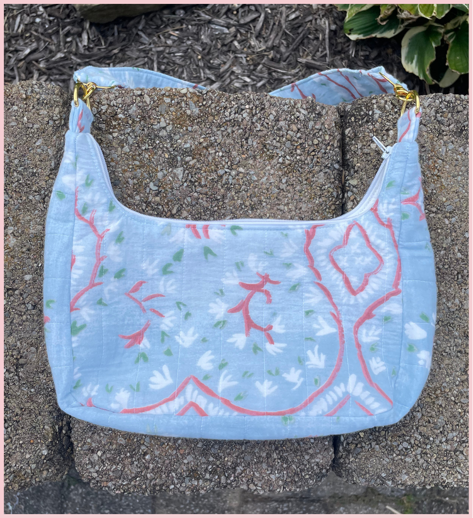 A light blue quilted fabric shoulder bag with a white floral pattern and coral-red botanical outlines, resting on gray concrete pavers outdoors.