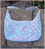 A light blue quilted fabric shoulder bag with a white floral pattern and coral-red botanical outlines, resting on gray concrete pavers outdoors.