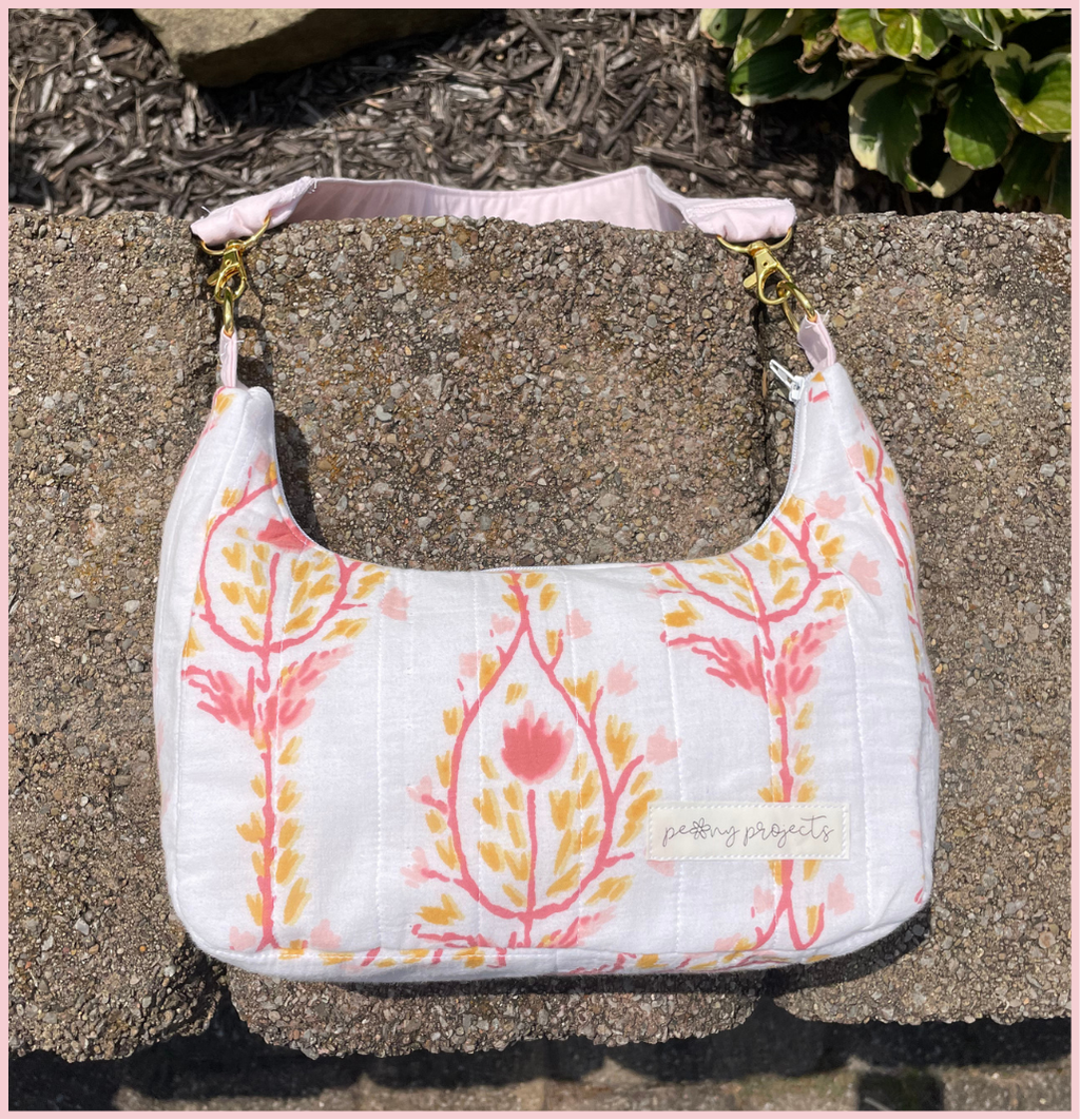 A white quilted fabric shoulder bag with a pink and orange botanical pattern, resting on a textured concrete surface outdoors.