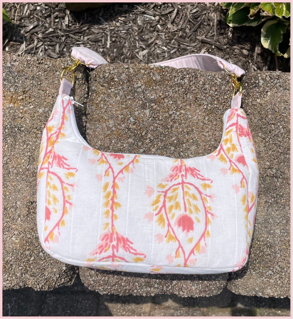 A white shoulder bag with a pink and yellow botanical pattern, resting on a textured concrete surface outdoors.