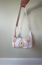 A hand holding a white fabric shoulder bag with a pink floral pattern and a pink strap, shown against a plain beige wall.