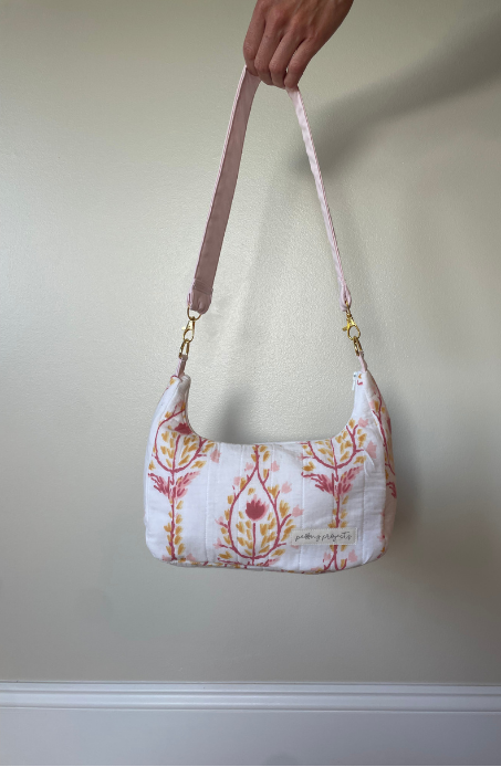 A hand holding a white fabric shoulder bag with a pink floral pattern and a pink strap, shown against a plain beige wall.