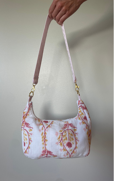 A white shoulder bag with a pink and yellow botanical pattern, held by a hand against a plain light gray wall.