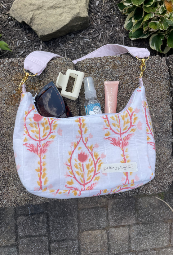 A white fabric shoulder bag with a pink and orange floral tree pattern, shown outdoors on a stone surface with its strap laid across it. The bag contains sunglasses, a hair clip, a spray bottle, and a pink tube.