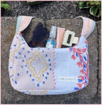 A pastel patchwork fabric shoulder bag with blue, pink, and yellow patterns, shown outdoors on a stone surface with sunglasses, a hand sanitizer bottle, a pink tube, and a cream hair clip inside.