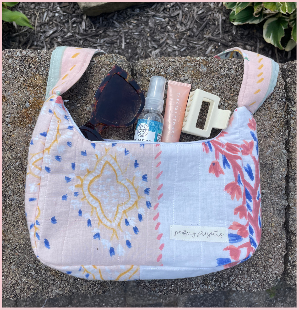 A pastel patchwork fabric shoulder bag with blue, pink, and yellow patterns, shown outdoors on a stone surface with sunglasses, a hand sanitizer bottle, a pink tube, and a cream hair clip inside.