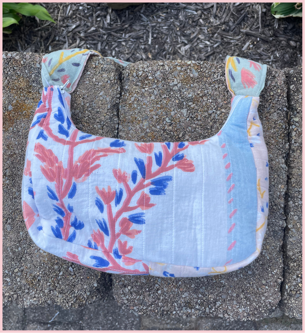 A small shoulder bag made from light blue crinkled fabric with a coral-pink and blue botanical leaf pattern, resting on a gray stone surface outdoors.