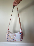 A handmade quilted shoulder bag with a floral and geometric patchwork pattern, held by a hand against a plain beige wall.