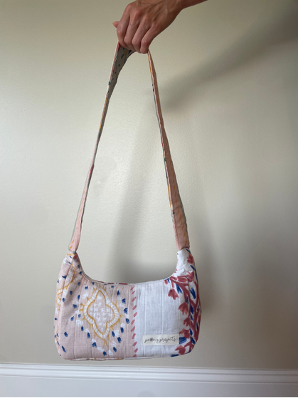 A handmade quilted shoulder bag with a floral and geometric patchwork pattern, held by a hand against a plain beige wall.