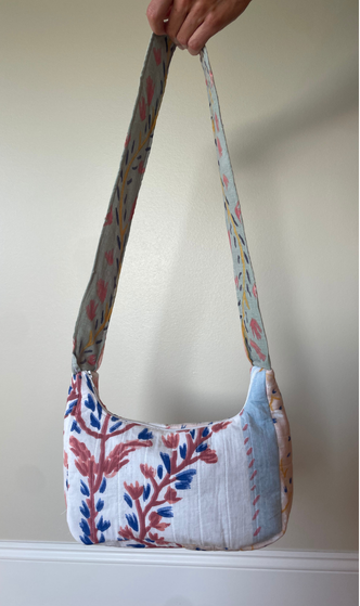 A handmade shoulder bag with a floral patterned fabric, held by a hand against a plain beige wall.