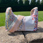 A handmade muslin baby bag with a floral and geometric pattern in pink, blue, yellow, and green on a white base, resting on a stone wall with a green grassy background.