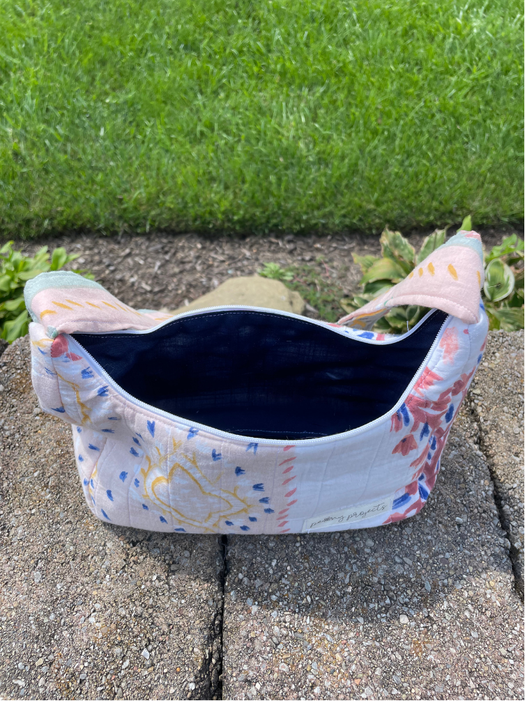 A small fabric pouch or cosmetic bag with a white base, colorful floral and geometric print in pink, blue, and yellow, and a dark navy blue interior, sitting outdoors on a stone surface with grass in the background.
