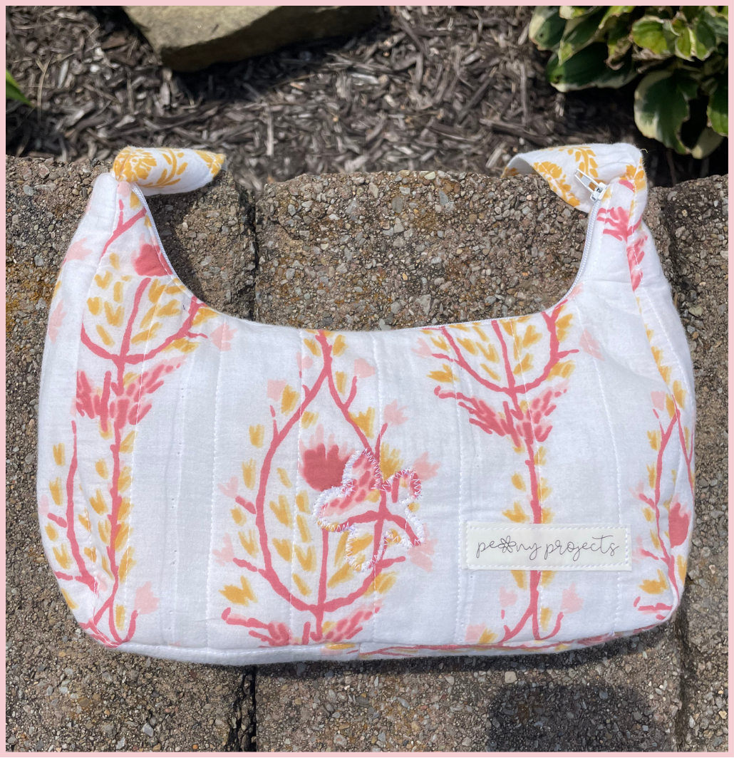 A small fabric shoulder bag with a white background and a pink and yellow botanical pattern, resting on a stone surface outdoors. A small fabric label on the front reads 'petty projects'.