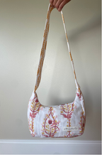 A fabric shoulder bag with a white base and a pink and orange botanical pattern, held by a hand against a plain beige wall.