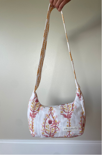 A fabric shoulder bag with a white base and a pink and orange botanical pattern, held by a hand against a plain beige wall.
