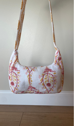 A small shoulder bag with a white base and a pink and yellow botanical pattern, hanging against a beige wall above a white baseboard and light wood floor.