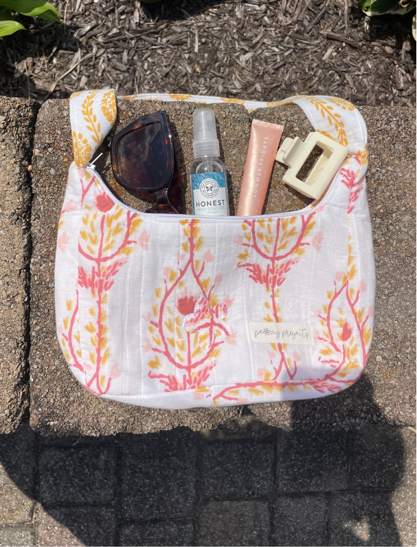 A white muslin shoulder bag with a pink and orange botanical pattern, shown outdoors on a stone surface with sunglasses, a spray bottle, a tube, and a hair clip inside.