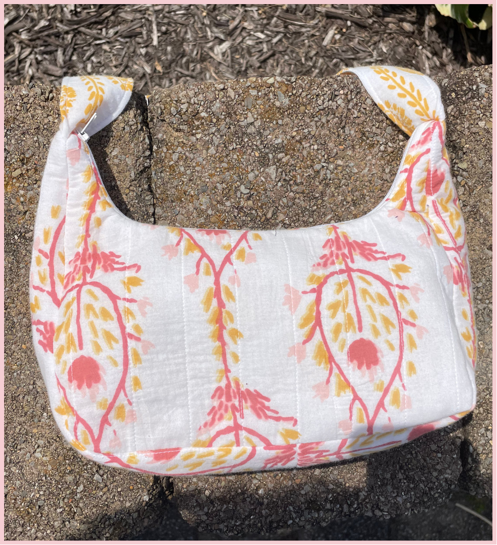 A white shoulder bag with a pink and yellow botanical pattern, photographed outdoors on a concrete surface with mulch in the background.