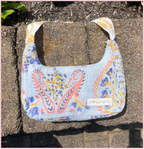A light blue fabric shoulder bag with a colorful paisley and floral pattern, resting on concrete pavers outdoors. The bag has a white shoulder strap and a small white label that reads 'patching projects'.