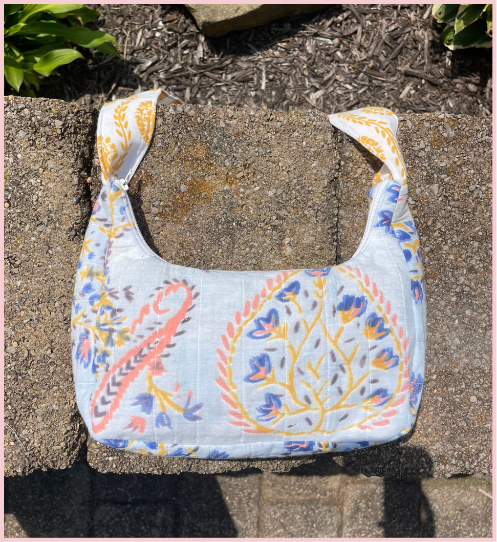 A fabric shoulder bag with a light blue background and a colorful floral and paisley print, resting on a concrete block outdoors.