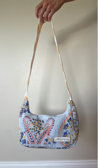 A hand holding up a small shoulder bag with a light blue base and a colorful paisley pattern in pink, yellow, and blue. The bag has a matching patterned strap and a small white label that reads 'picking pretty'.