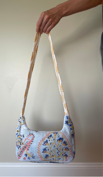 A hand holding a small fabric shoulder bag with a light blue background and a multicolor floral and paisley print, featuring a long patterned strap.