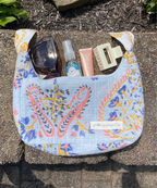 A light blue fabric shoulder bag with a colorful paisley pattern, shown outdoors on stone pavers with sunglasses, a hand sanitizer bottle, a pink tube, and a cream hair clip inside the pocket. A small white label on the front reads 'picking projects'.
