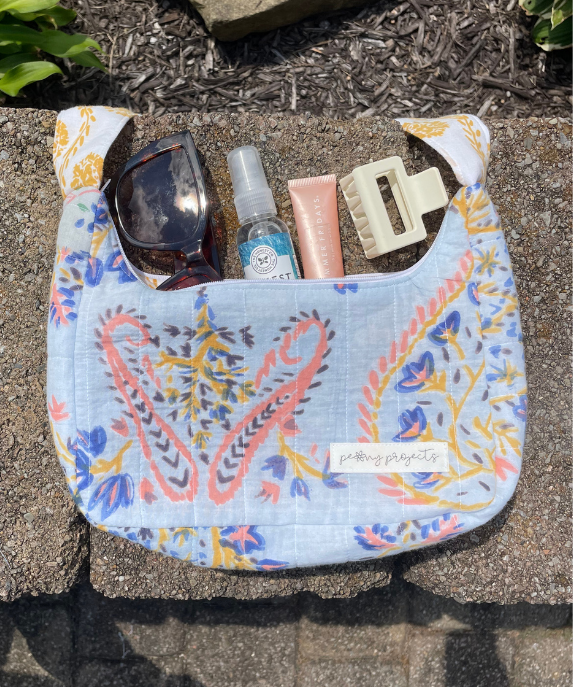 A light blue fabric shoulder bag with a colorful paisley pattern, shown outdoors on stone pavers with sunglasses, a hand sanitizer bottle, a pink tube, and a cream hair clip inside the pocket. A small white label on the front reads 'picking projects'.
