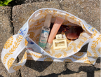A white tote bag with a yellow botanical pattern is shown outdoors on stone pavers, filled with various personal care items including a bottle of hand sanitizer, a pink tube, and sunglasses.