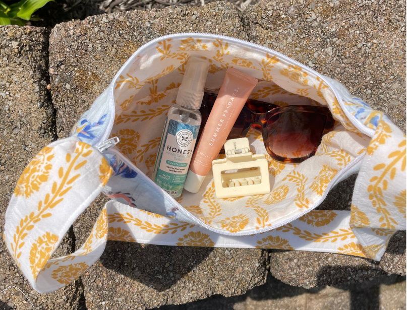 A white tote bag with a yellow botanical pattern is shown outdoors on stone pavers, filled with various personal care items including a bottle of hand sanitizer, a pink tube, and sunglasses.