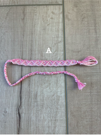 A pink and white braided friendship bracelet with a tassel, laid flat on a gray wood plank background. A small white letter 'A' is printed near the center.