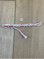 A handmade braided friendship bracelet with a colorful geometric pattern in pink, green, yellow, and white, laid flat on a light brown wooden surface. A white text overlay reads 'D'.