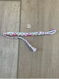 A handmade braided friendship bracelet with a colorful geometric pattern in pink, green, yellow, and white, laid flat on a light brown wooden surface. A white text overlay reads 'D'.