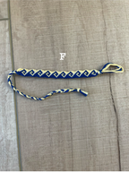 A blue and white braided rope accessory with a gold metal clasp, laid flat on a textured gray fabric background.