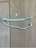 A handmade braided friendship bracelet in light blue and yellow, laid out on a brown wood-grain floor. The word 'E' is printed above the bracelet.