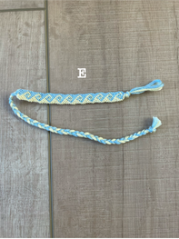 A handmade braided friendship bracelet in light blue and yellow, laid out on a brown wood-grain floor. The word 'E' is printed above the bracelet.
