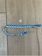 A blue and white braided friendship bracelet with a geometric pattern, laid flat on a light brown wood background. A white letter 'H' is overlaid near the center.