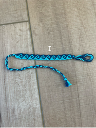 A handmade friendship bracelet made of bright blue and black braided cord, laid out on a light brown wood floor. A white label with the letter 'I' is placed near the center of the bracelet.