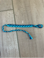 A handmade friendship bracelet made of bright blue and black braided cord, laid out on a light brown wood floor. A white label with the letter 'I' is placed near the center of the bracelet.