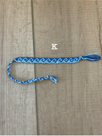 A blue and white braided friendship bracelet with a tassel, laid flat on a light wood background. The letter 'K' is printed above the bracelet.