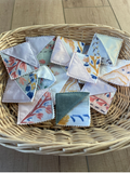 A collection of small square fabric napkins with various floral and abstract prints, arranged inside a round woven wicker basket on a light wooden floor.