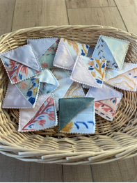 A collection of small square fabric napkins with various floral and abstract prints, arranged inside a round woven wicker basket on a light wooden floor.