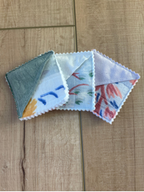 Three small square fabric napkins with scalloped edges fanned out on a light brown wooden surface. The napkins feature different pastel watercolor designs, including blue, red, pink, and yellow motifs on white, gray, and light purple backgrounds.