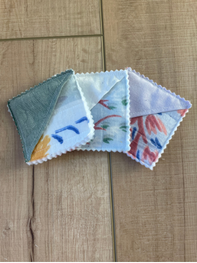 Three small square fabric napkins with scalloped edges fanned out on a light brown wooden surface. The napkins feature different pastel watercolor designs, including blue, red, pink, and yellow motifs on white, gray, and light purple backgrounds.