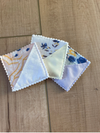 Three small triangular fabric napkins with scalloped edges are arranged on a light brown wooden surface. The napkins feature delicate floral and geometric patterns in soft pastel colors including blue, yellow, and white.