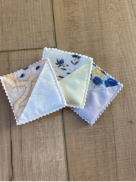Three small triangular fabric napkins with scalloped edges are arranged on a light brown wooden surface. The napkins feature delicate floral and geometric patterns in soft pastel colors including blue, yellow, and white.
