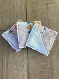 Four square cotton handkerchiefs with scalloped edges fanned out on a light brown wooden surface. The fabrics feature various prints, including solid white, light blue, and marbled orange and white patterns.