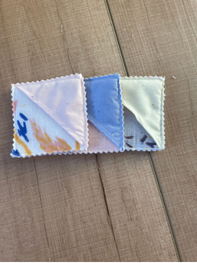 Three small square fabric napkins or handkerchiefs with scalloped edges laid out on a wooden surface. The left napkin is white with a blue and yellow abstract print, the middle napkin is light blue, and the right napkin is pale green with dark blue abstract print.