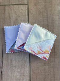 Three small square fabric items with scalloped edges, fanned out on a light wood surface. The fabrics appear in soft pastel colors, including pale blue, white, and a very light mint or green with faint floral accents.