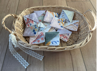 A wicker basket with handles sits on a light wood floor against a neutral wall, holding several small folded fabric pieces with colorful printed designs and white ribbon ties.